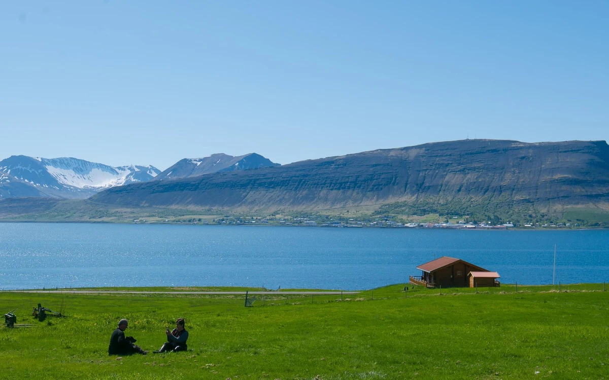 Two people sitting on grass above the fjord, with the town and flat-topped mountains across the water
