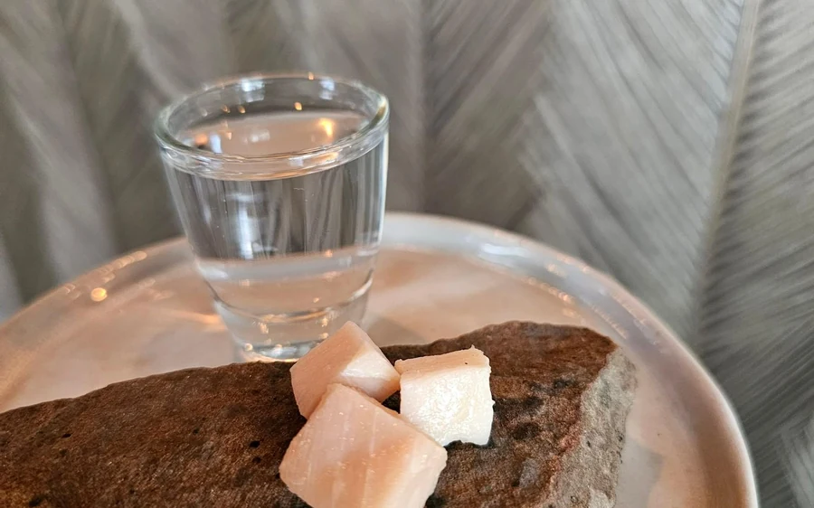 A shot glass beside dark rye bread and cubes of dried fish